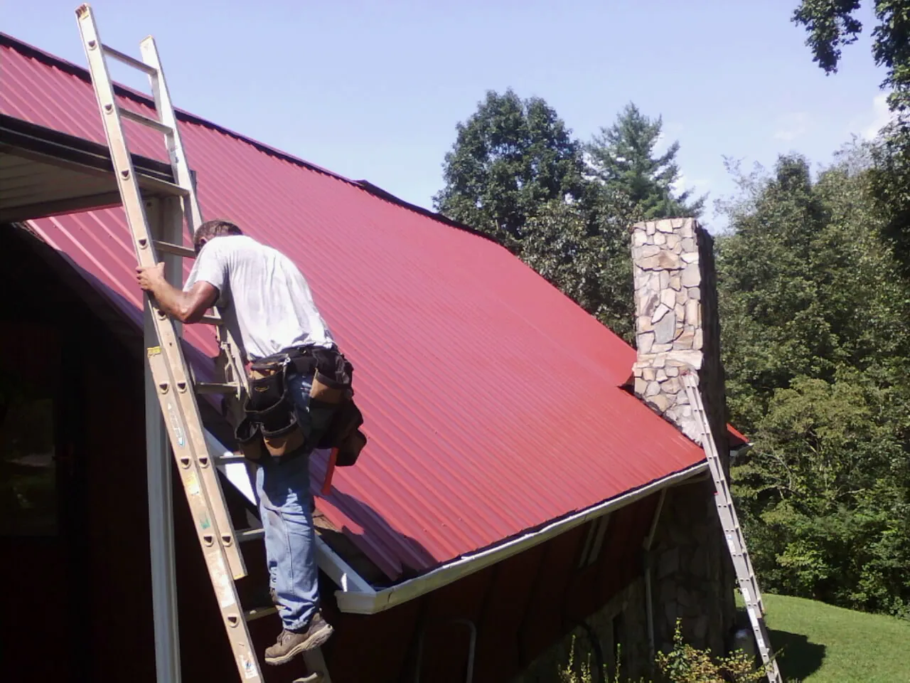 Roofer on a ladder beside a red metal roof and stone chimney — Soffit & Fascia Repair by Roofing Brothers in Huson